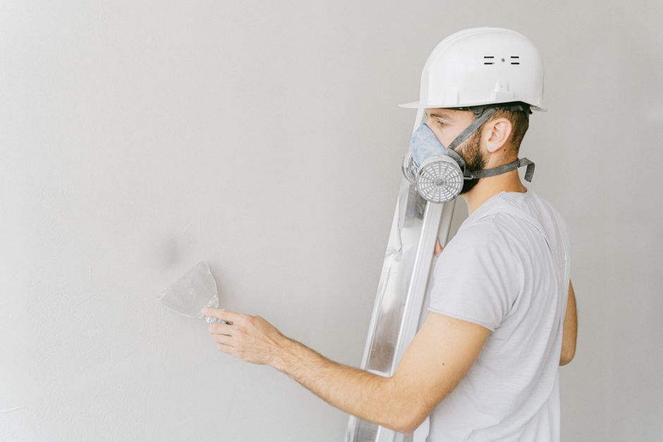 A construction worker wearing a gas mask and hard hat applies plaster to a wall indoors.