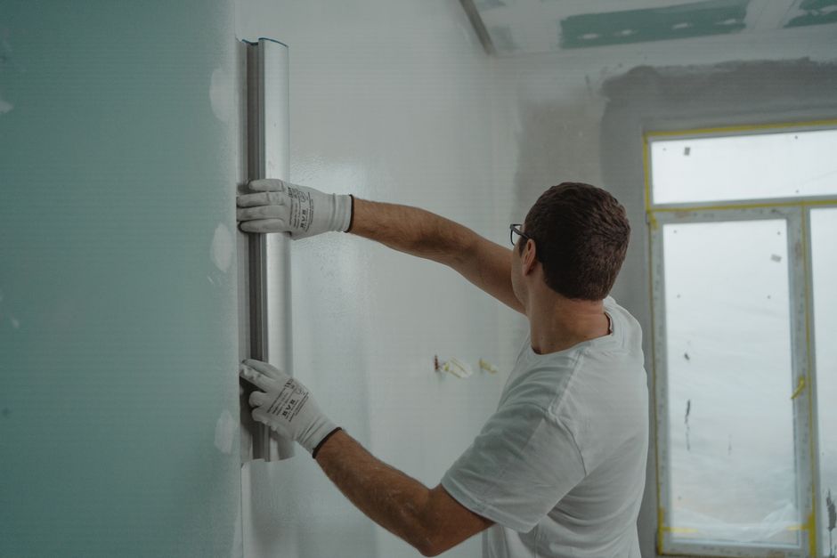 A man in a white shirt and gloves using a metal ruler for indoor wall renovation work.